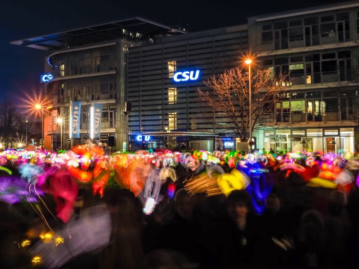 Tausende malen mit farbigen Lichtern in der Dunkelheit vor der CSU Parteizentrale in München.